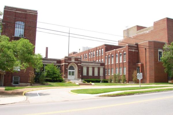 The image shows a large brick building with multiple sections, surrounded by grass and trees, located along a street with a sidewalk.