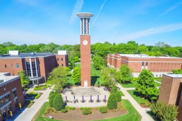 An aerial view of a campus with a brick clock tower, surrounded by multiple buildings and greenery under a clear blue sky.