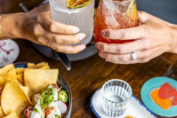 Two hands hold drinks over a table with chips, ceviche, and a tray of sliced mango. Coasters are visible underneath the drinks.