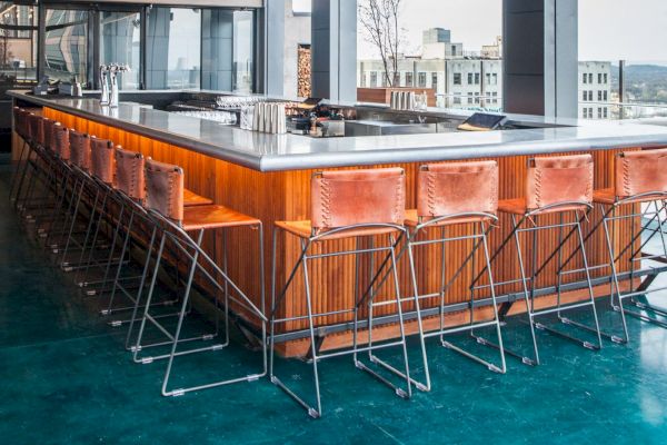 Modern bar with a sleek countertop, surrounded by stylish metal and leather bar stools, located in a high-rise with large windows.