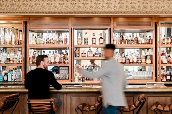 A stylish bar with bottles displayed, a seated person, and another walking by, surrounded by elegant wooden and metal decor.