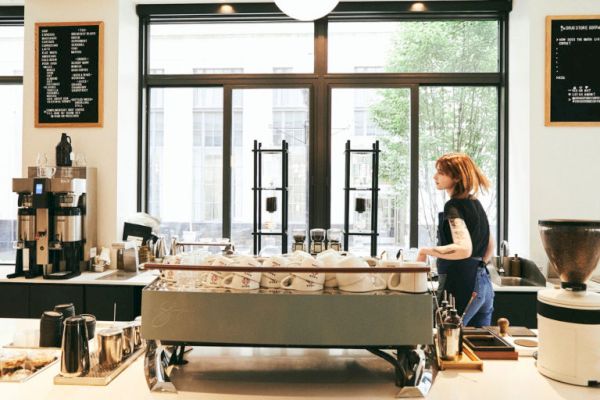 A barista is working in a modern coffee shop with large windows and menus on the wall, surrounded by coffee equipment and decor.