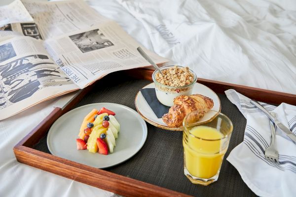 A breakfast tray with fruit, a croissant, granola, orange juice, and a newspaper on a bed.