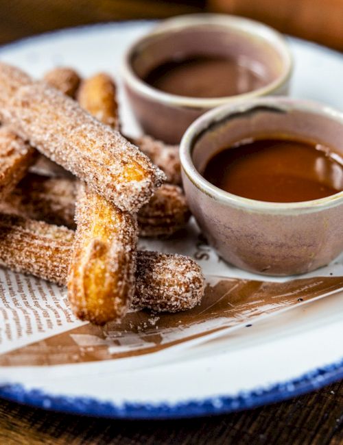 The image shows churros on a plate with two cups of chocolate dipping sauce.