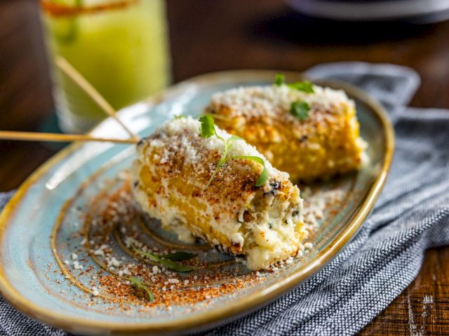 The image shows a plate with elote, a Mexican street corn dish, garnished with cheese and spices, set on a cloth with a drink in the background.