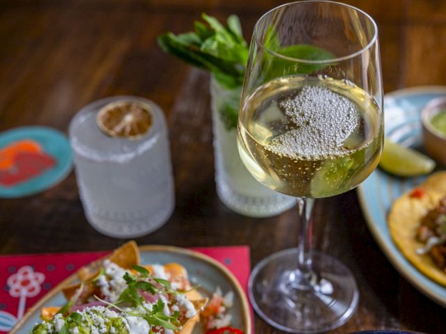 A glass of white wine, a cocktail, another drink with citrus garnish, and plates of food, likely tacos, on a wooden table.