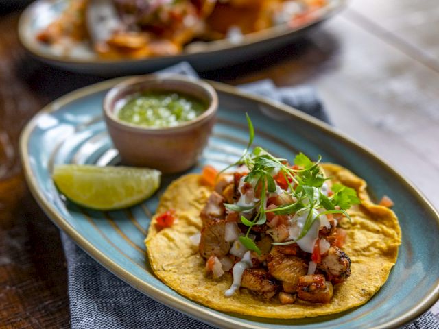 A taco with grilled meat, fresh greens, radish, and sauce is on a ceramic plate, served with a lime wedge and a small bowl of green sauce.