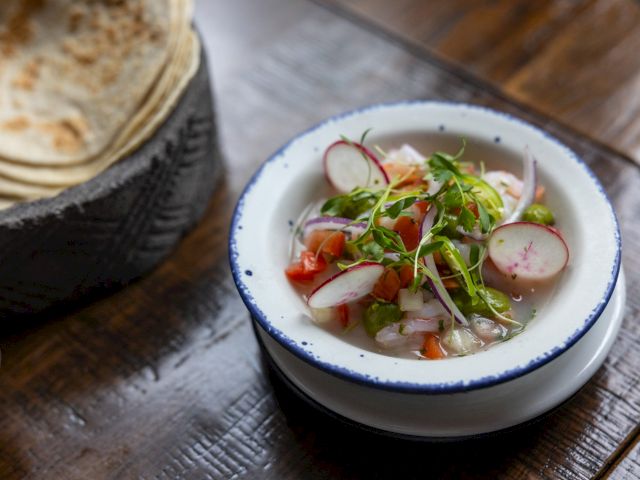 The image shows a bowl of ceviche with radish slices, herbs, and tomatoes, next to a basket of flatbreads on a wooden table.