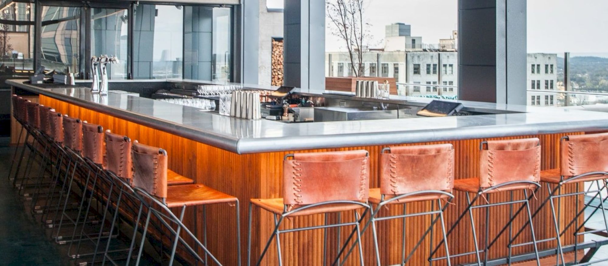 The image shows a modern bar setup with leather chairs and a sleek countertop, located in a bright, open space with a city view.