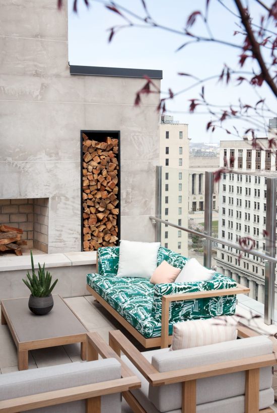 A rooftop patio with a sofa, cushions, coffee table, fireplace, and wood stack, overlooking a cityscape with buildings in the background.