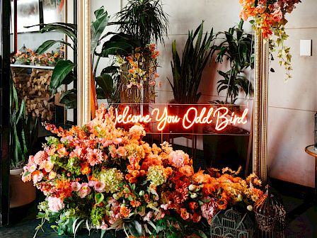 A floral arrangement with a large frame, neon sign, and assorted flowers; plants in the background enhance the decor.