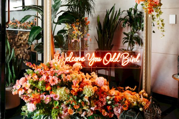 A floral arrangement with a large frame, neon sign, and assorted flowers; plants in the background enhance the decor.