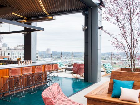 This image shows a modern rooftop bar with wooden furnishings, high stools, vibrant seating, and a city view in the background.