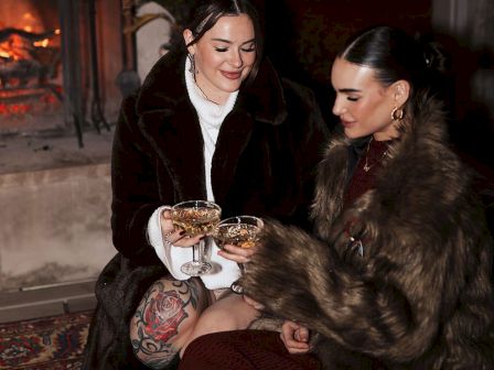 Two people wearing fur coats are sitting by a fireplace, holding drinks and smiling.