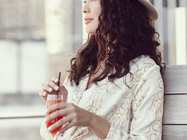 A person in a hat and patterned shirt is sitting, holding a red drink with a straw, looking to the side against an urban backdrop.