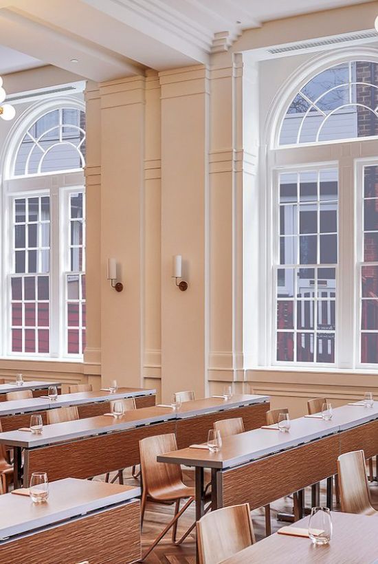 The image shows a spacious, well-lit classroom with rows of tables and chairs, large arched windows, and elegant lighting fixtures.