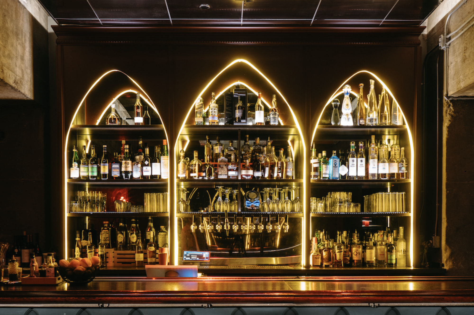 A dimly lit bar with illuminated shelves holding various bottles of alcohol and glassware, creating a warm, inviting atmosphere.