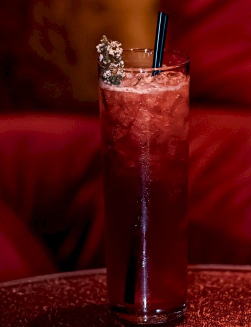 A tall glass with a red drink, ice, a straw, and a garnish sits on a table, against a red cushioned background.