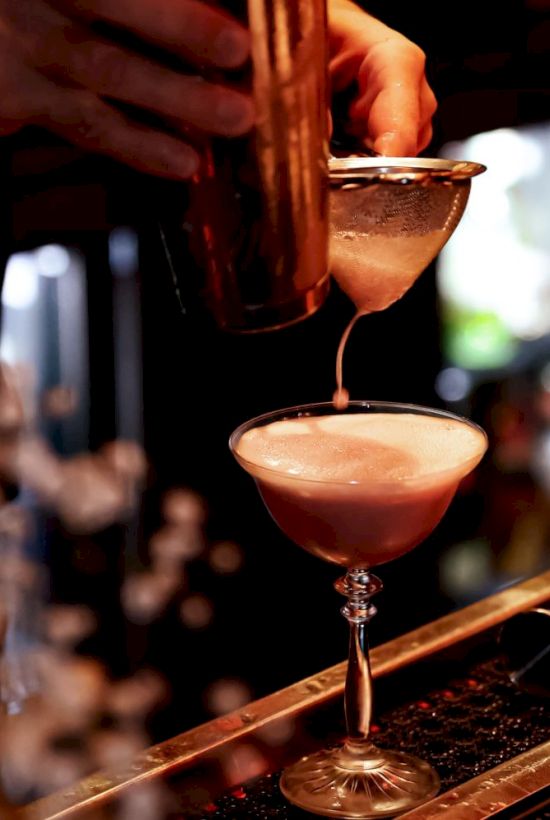 A bartender is pouring a cocktail through a strainer into a glass, set on a bar counter.