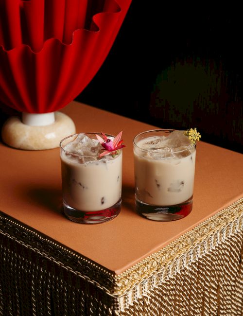 Two glasses of iced creamy beverages with garnishes sit on a table with a textured base and red fan-shaped decor in the background.