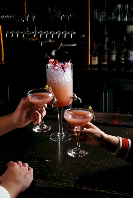 The image shows three people raising different pink cocktails, two in coupe glasses and one with crushed ice in an ornate glass.