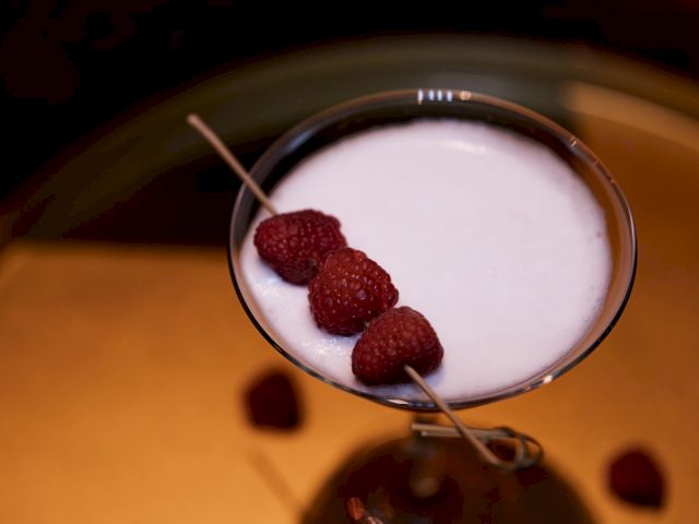 A creamy cocktail garnished with raspberries on a skewer, served in a martini glass.