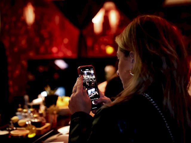 A person is taking a photo with a smartphone in a dimly lit, possibly restaurant setting, with drinks and dim lights visible in the background.