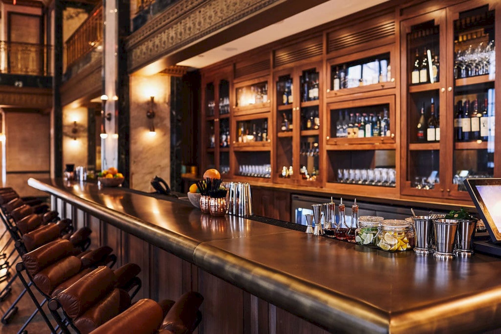 A stylish bar with wooden shelves, wine bottles, glasses, bar stools, and various cocktail ingredients on the counter.