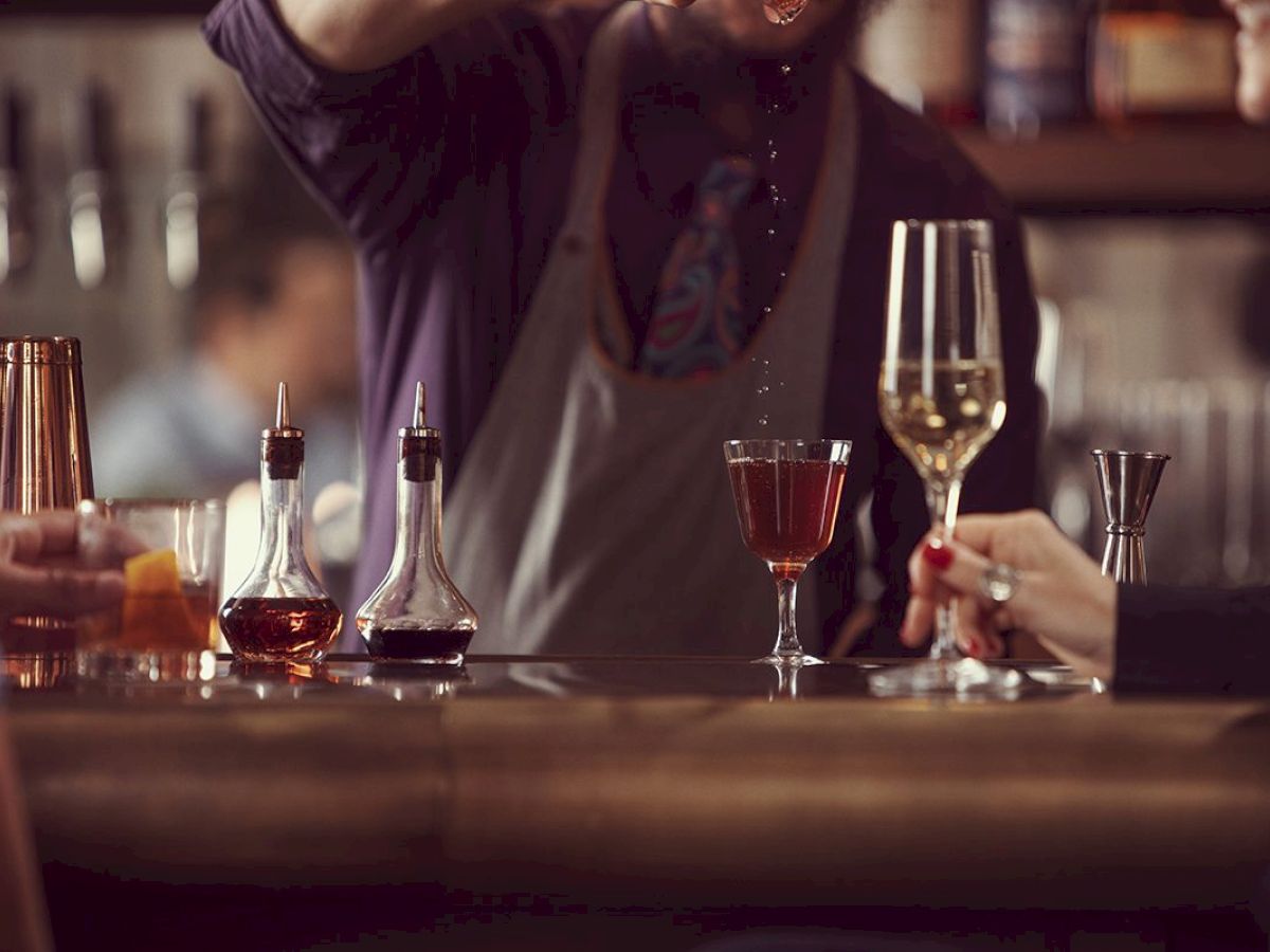 A bartender pours a drink at a bar. Two patrons hold glasses, surrounded by cocktail tools and ingredients, creating a cozy atmosphere.