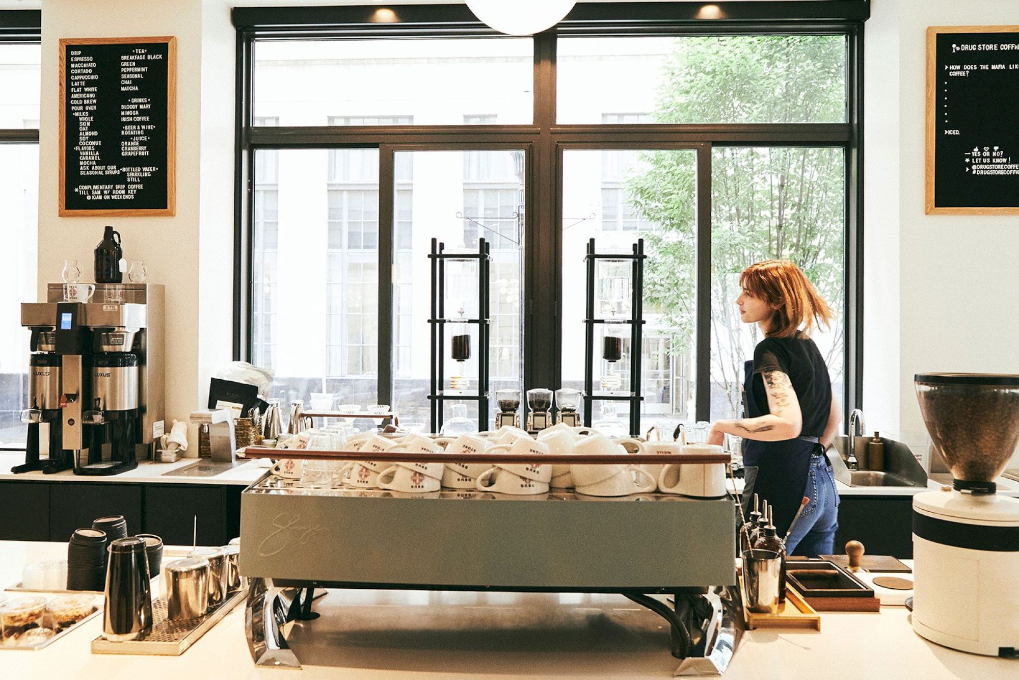 A barista working in a coffee shop with modern equipment, large windows, and a menu board visible on the wall.