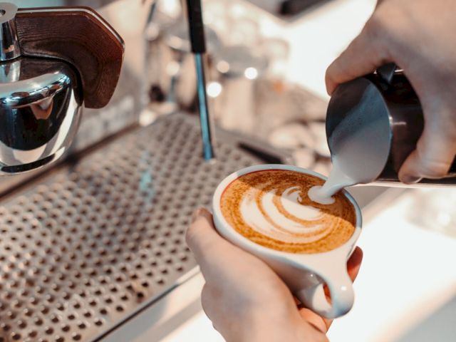 A person is pouring milk into a cup of coffee, creating latte art, next to an espresso machine in a cafe setting.