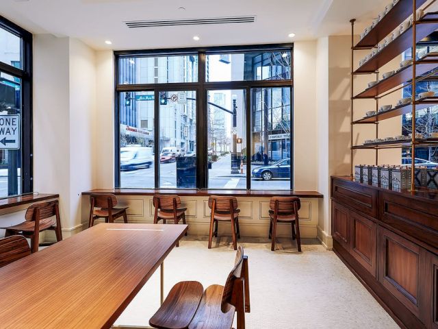 The image shows an empty, modern cafe interior with wooden tables and chairs, large windows, street view, and shelving on the right side.