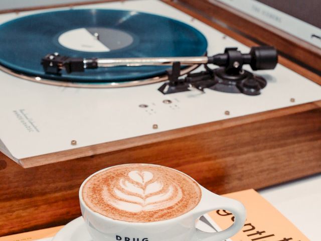 A latte with latte art in a cup on a saucer, placed on a magazine, with a vinyl record on a turntable in the background.