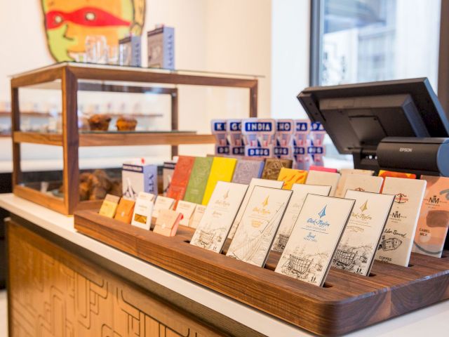 A countertop display with various chocolate bars, a pastry case in the background, and a cash register nearby.