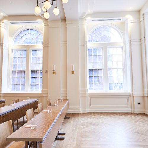 A bright room with arched windows, wooden floors, and rows of tables and chairs. Modern lighting complements the elegant design.