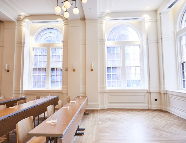 A bright room with arched windows, wooden floors, and rows of tables and chairs. Modern lighting complements the elegant design.