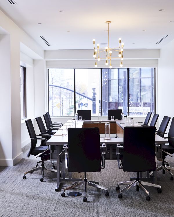 A modern conference room with a long table, office chairs, large windows, and a chandelier, offering a bright office setting.