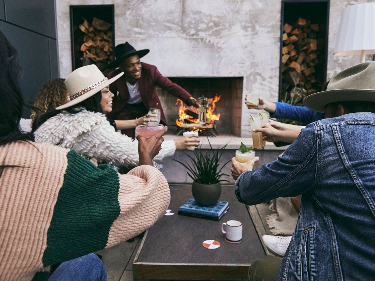 A group of people wearing casual outfits and hats enjoy drinks around a fire, in a cozy setting with an outdoor fireplace.