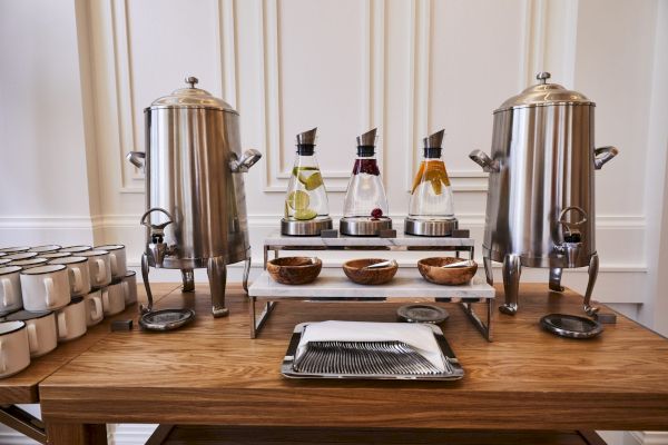 The image shows a beverage station with dispensers, infused water, bowls, mugs, and a tray on a wooden table against a white wall.