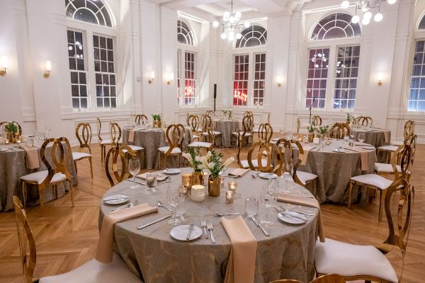 An elegantly set banquet hall with round tables, decorative candles, and arranged cutlery, featuring large windows and sophisticated lighting.