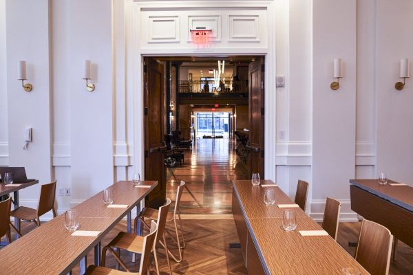 A dining area with wooden tables and chairs; two glasses per table, leading to a hallway and a window view at the end of the room.