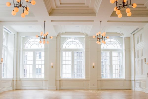 The image shows an elegant, empty room with large windows, chandeliers, and detailed molding on the walls and ceiling.