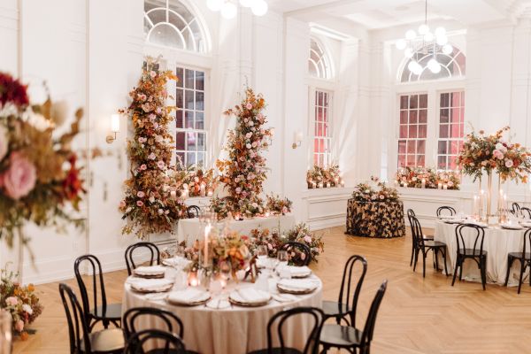 Elegant event setup with round tables, floral decorations, and candles in a bright, white room with arched windows and wooden floor.