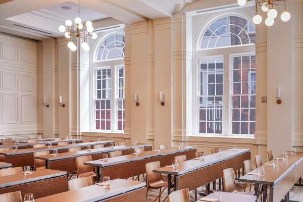 The image shows a stylish, well-lit conference room or classroom with long tables, chairs, and large arched windows lining the walls.