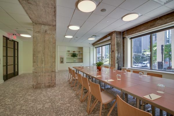 A modern conference room with a long table, wooden chairs, potted plant, and large windows overlooking a cityscape.