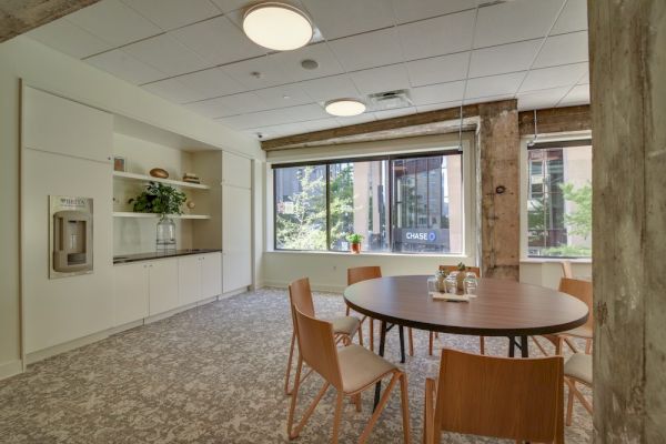 The image shows a modern office break room with a round table, chairs, a window view, and built-in cabinets with a water dispenser.