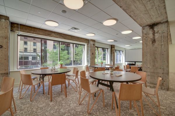 A modern dining area with round tables, wooden chairs, large windows, and exposed concrete pillars.