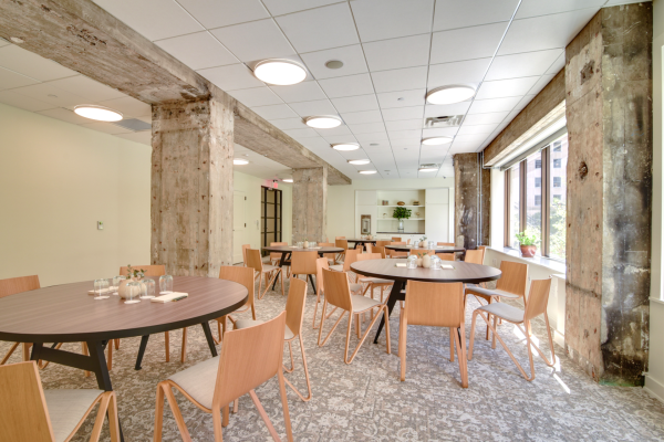 The image shows a modern, well-lit dining area with wooden tables, chairs, and large windows, featuring exposed concrete beams.