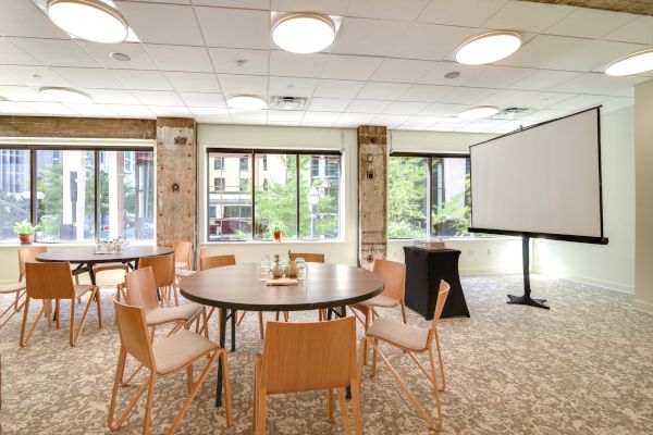 A conference room with round tables, chairs, large windows, and a presentation screen. Urban view outside.