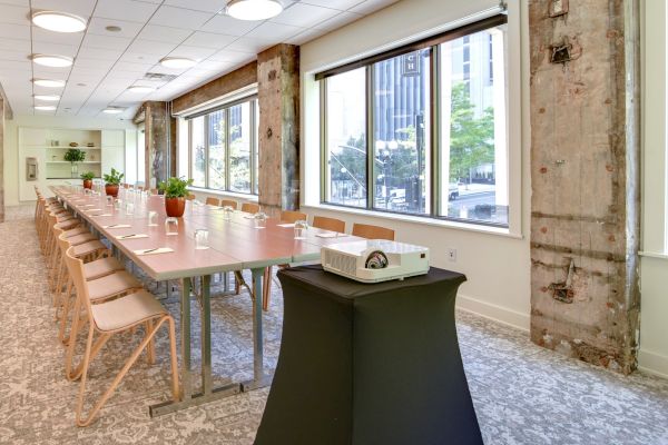 A long conference table with chairs and a projector in a well-lit room with large windows and exposed beams ends the sentence.